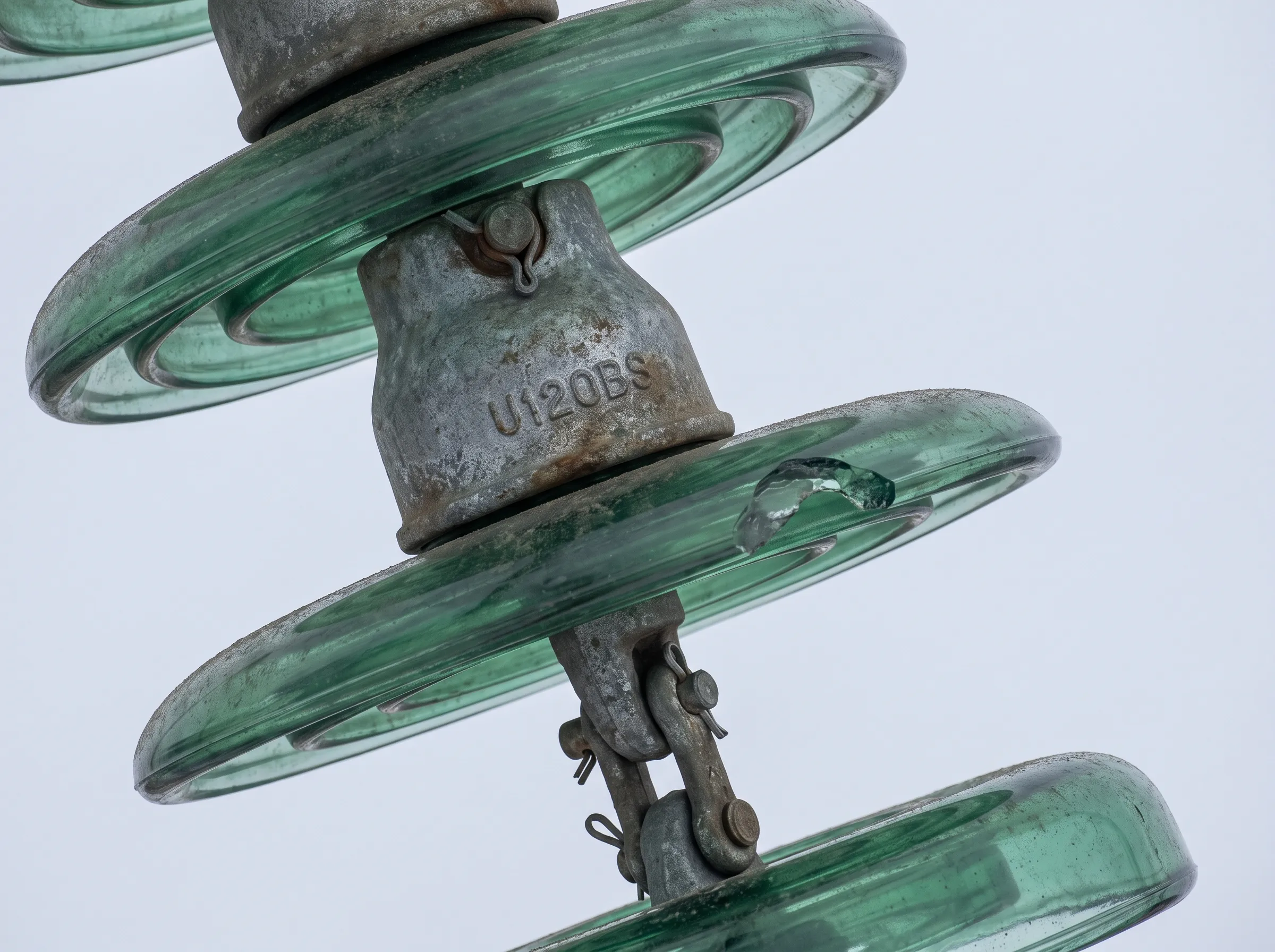 Gros plan d'un isolateur : la référence U120BS frappée sur le capot d'acier galvanisé, et un éclat sur le bord de l'assiette de verre.